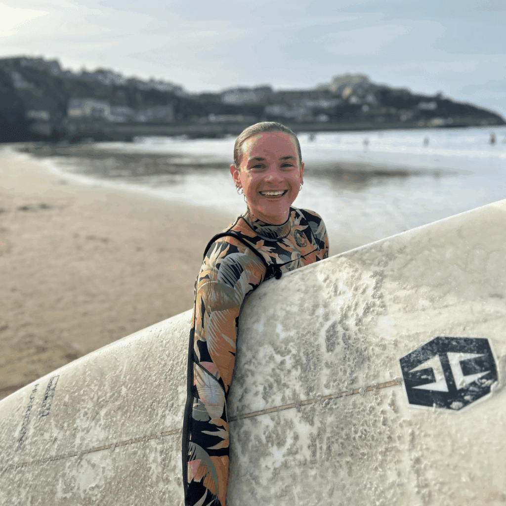 Sophie Ellis with wet hair, stood on the beach in a wetsuit holding her surfboard.
