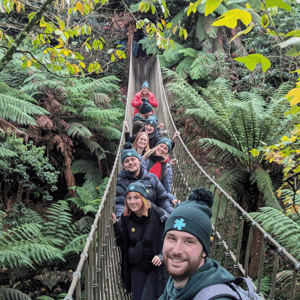 Hiyield team members in a line crossing a rope bridge, smiling at the camera wearing Hiyield bobble hats.
