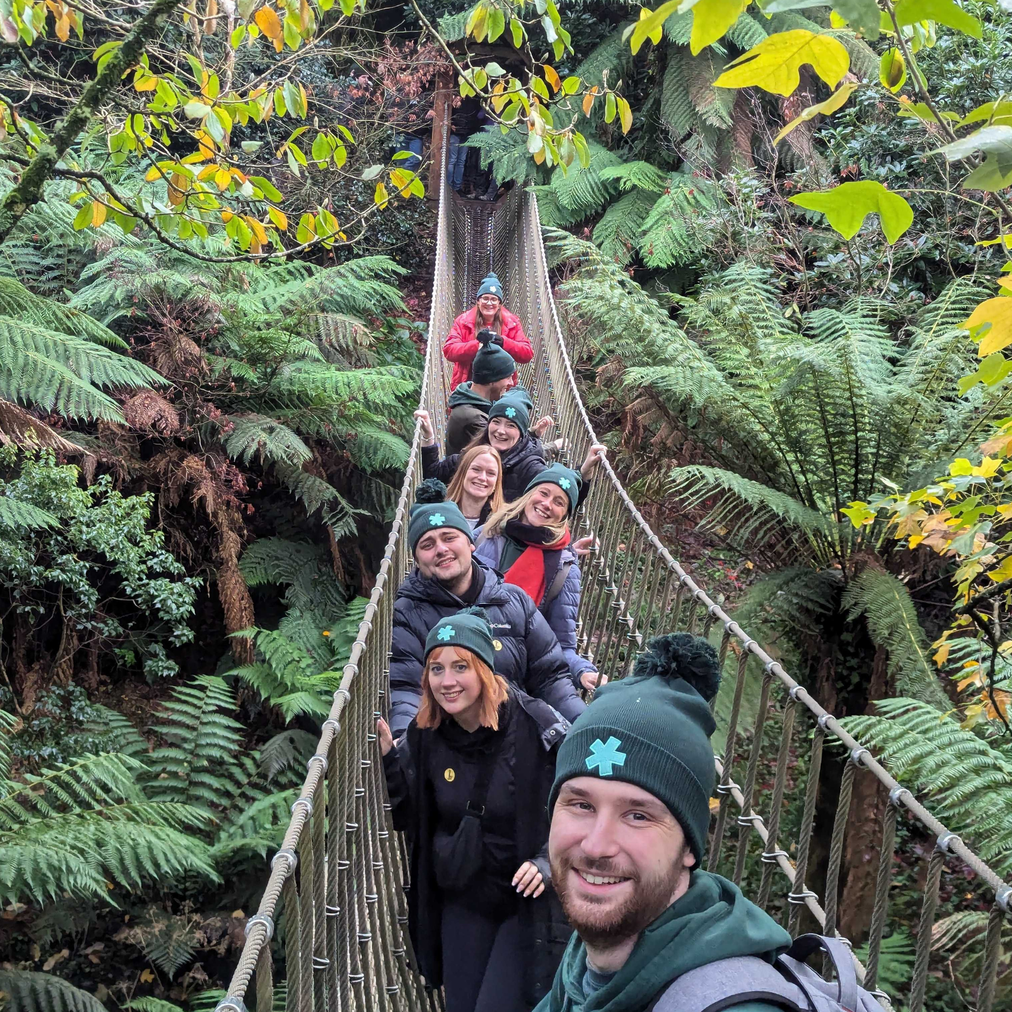 Hiyield team members in a line crossing a rope bridge, smiling at the camera wearing Hiyield bobble hats.