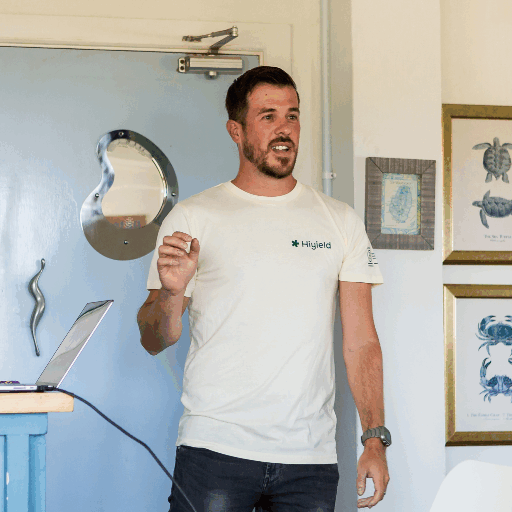 Matt Ville wearing a cream Hiyield t-shirt speaking the the team infront of a pale blue door in a nautical room.