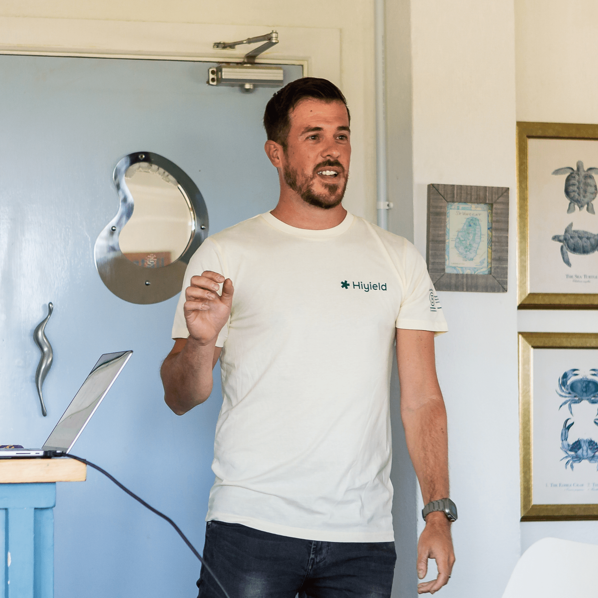 Matt Ville wearing a cream Hiyield t-shirt speaking the the team infront of a pale blue door in a nautical room.