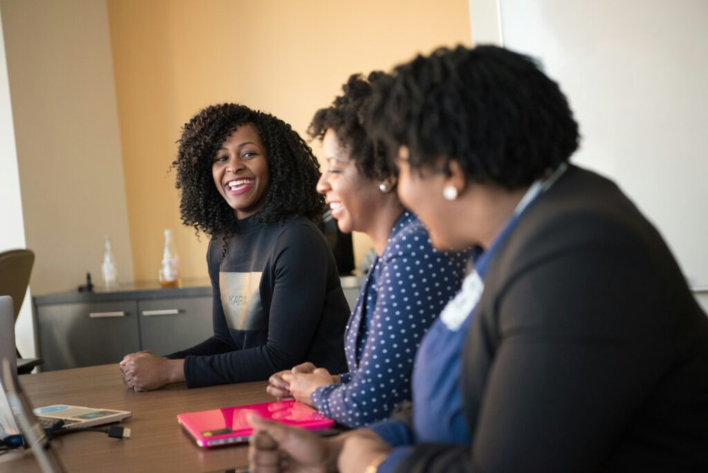 Three black women sat at a desk smiling during a business meeting.