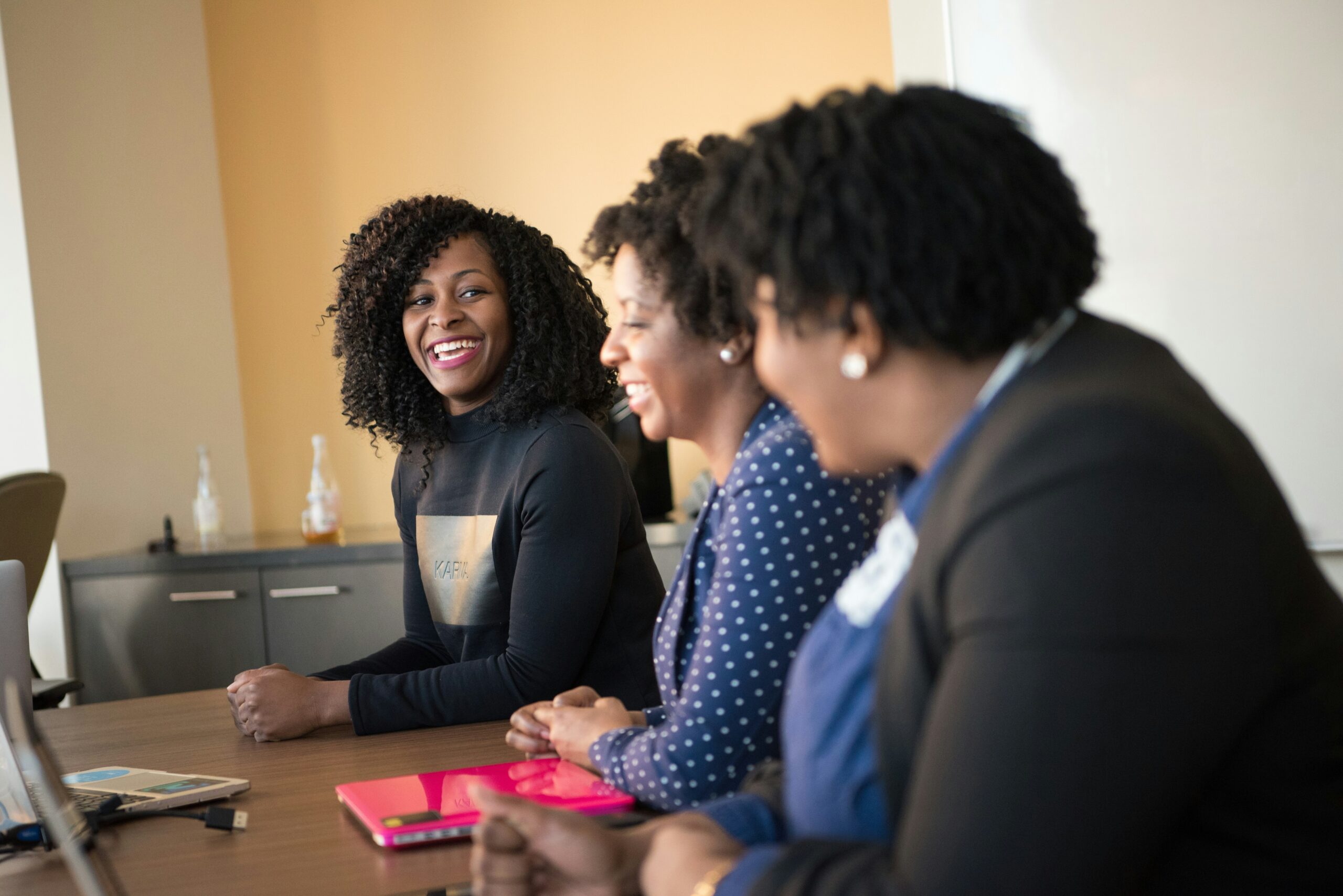 Three black women sat at a desk smiling during a business meeting.