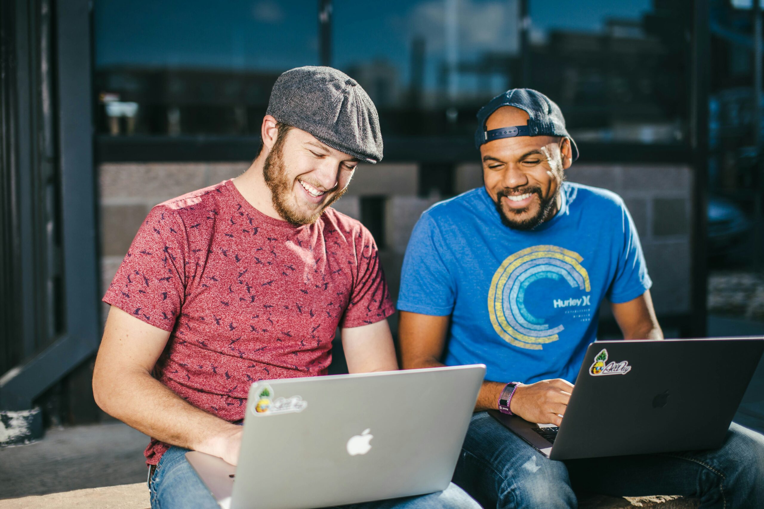 Two men in t-shirts smiling and looking at their laptops while sitting outside.