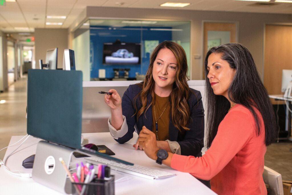 Two women are sat looking at a computer together, one woman is older with greying hair, the other is slightly younger with brown hair.