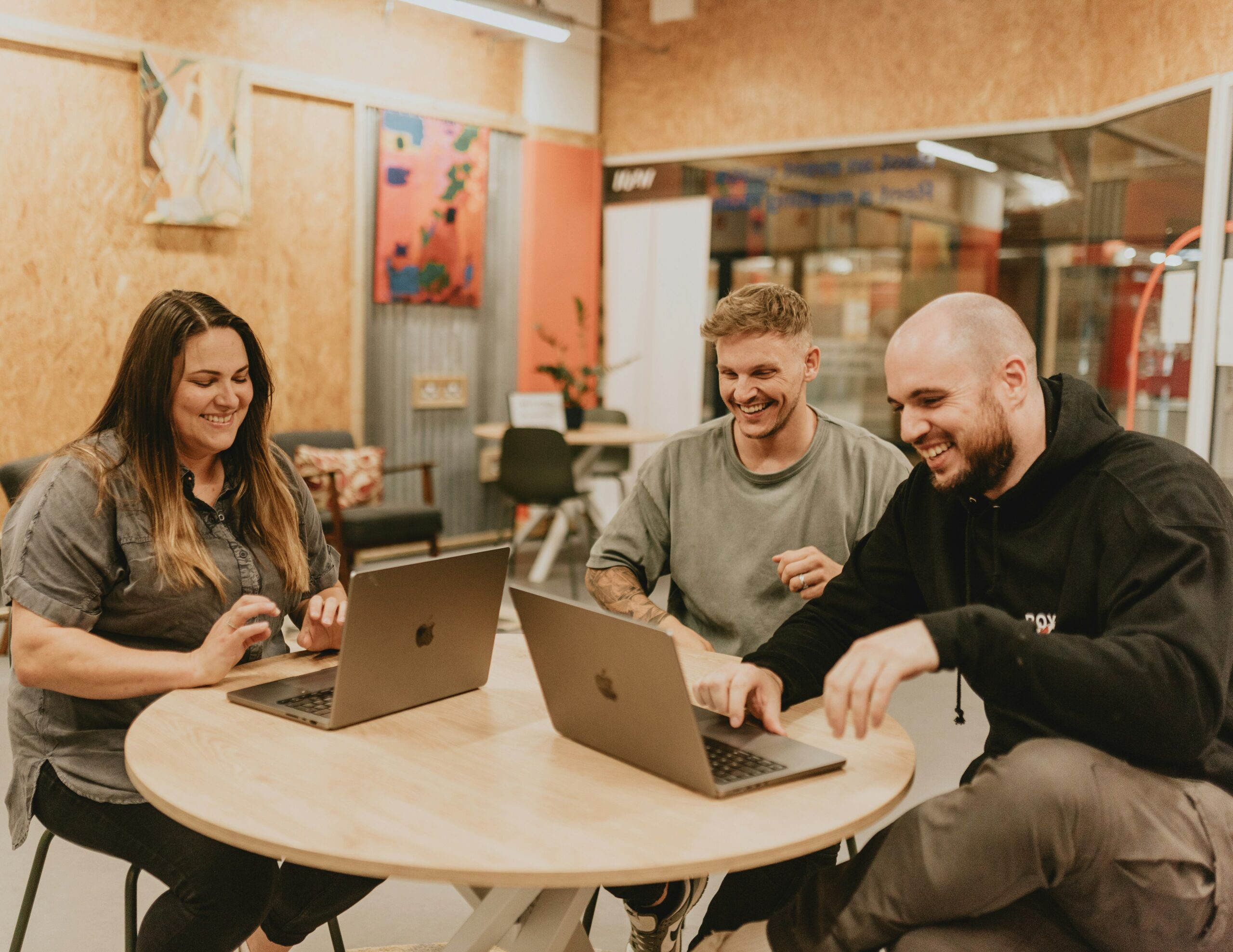 Two men and a woman sat at a wooden table with laptops, smiling at each other.