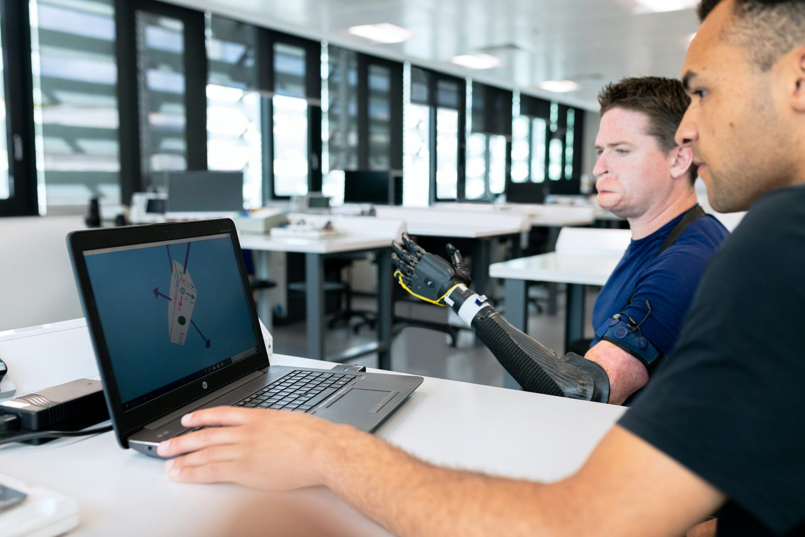 A male amputee using a wheelchair is getting a robotic arm fitted by another man.