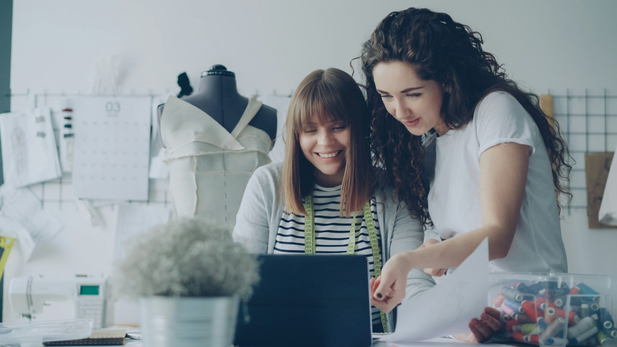 Two women smiling looking at a laptop, with textiles and sewing work in the background.
