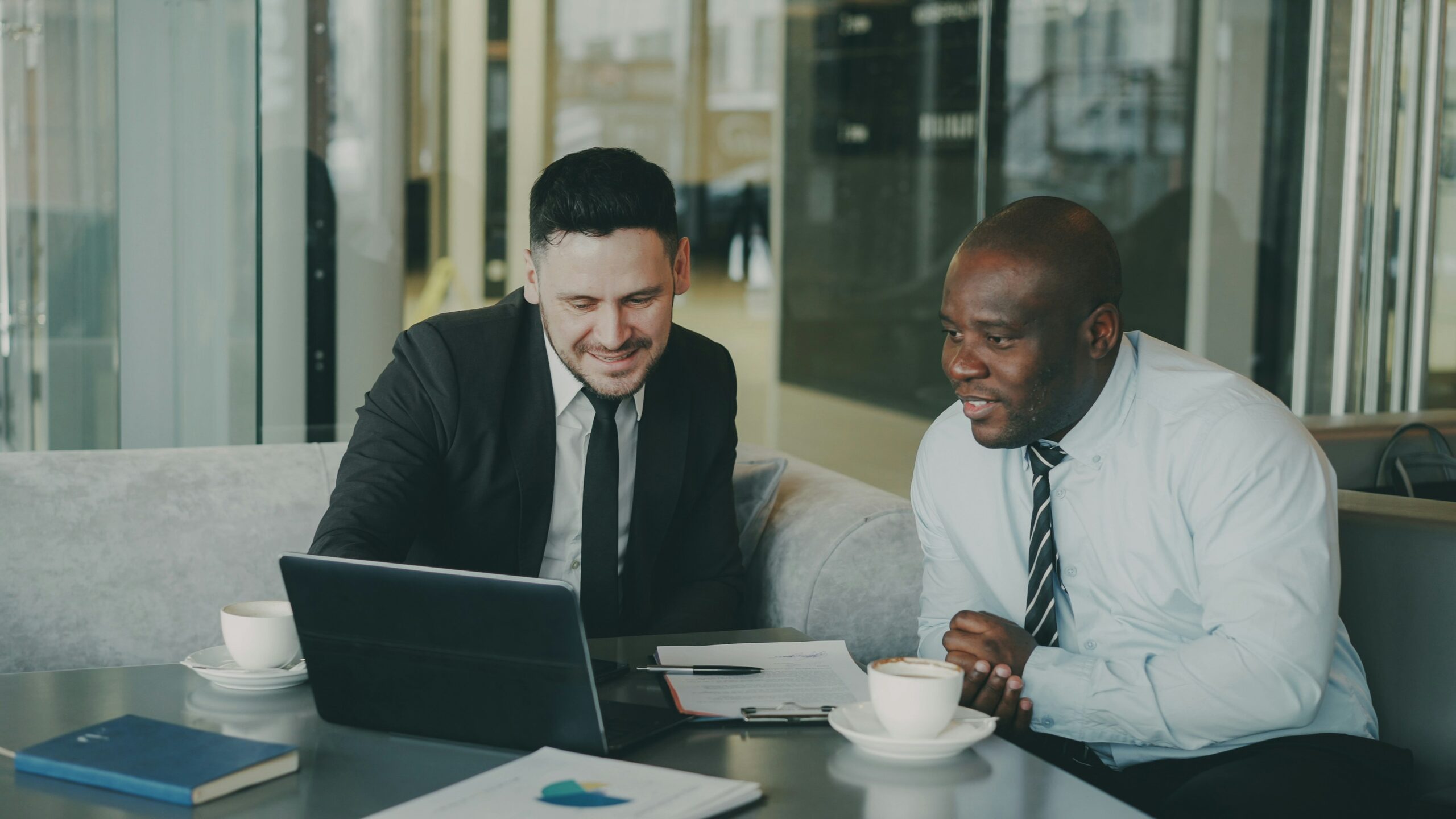 Two men in suits looking at their laptops on a table with coffee cups.