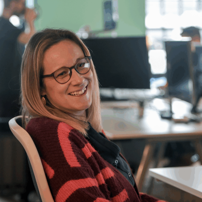 Photo of Laura Hudspith sat at her desk smiling, wearing a striped cardigan and glasses.
