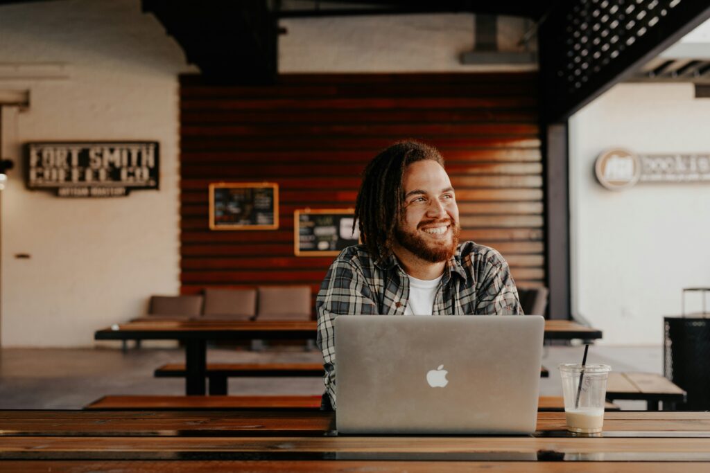 Man with long hair and beard sat at desk smiling with laptop with wooden panelling behind.