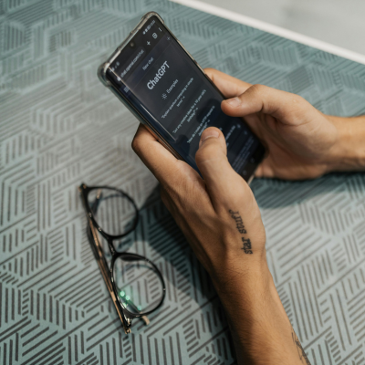 Hands holding a mobile phone with ChatGPT open, glasses sat on the table next to them.